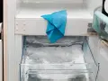 W zamrażarce lodówki, która głośno chodzi, zalega lód. Niebieska ściereczka wisi na krawędzi.