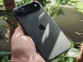 Ciemny iPhone z jednym aparatem w dłoni, otoczony zielenią roślin.