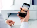 Dłoń trzyma smartfon z napisem "Sending to printer", pokazując jak drukować z telefonu. W tle biała drukarka.