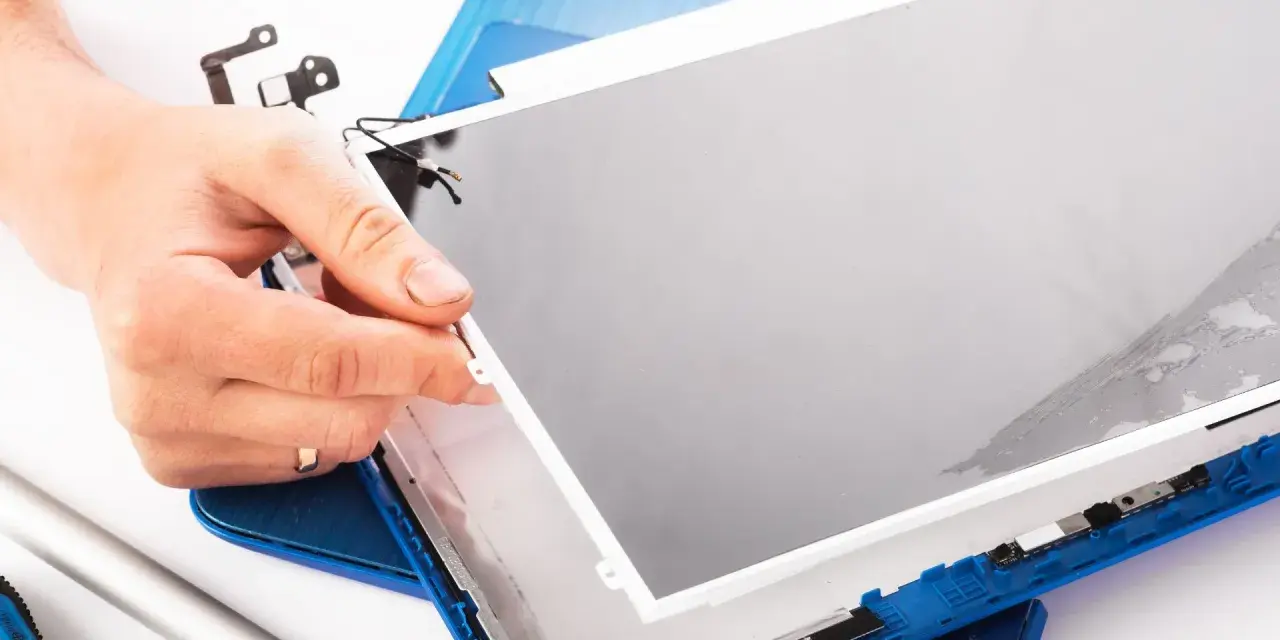 Dłoń trzyma matrycę w laptopie podczas naprawy. Widać narzędzia i niebieską obudowę.