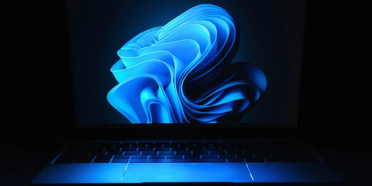 Niebieski, falujący kształt na ekranie laptopa z podświetlaną klawiaturą, przypominający logo Windows 11 24H2.