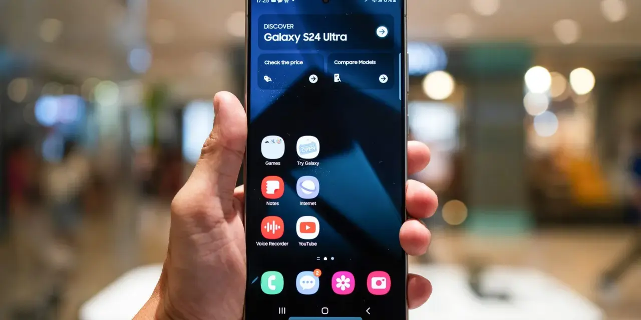 Dłoń trzyma smartfon z interfejsem One UI 6, prezentującym ekran startowy Galaxy S24 Ultra z ikonami aplikacji.