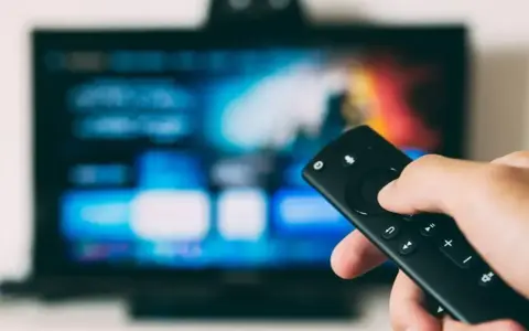 Dłoń trzymająca pilot do telewizora LG, ekran pokazuje menu. Dowiedz się, jak wyłączyć lektora w telewizorze LG.