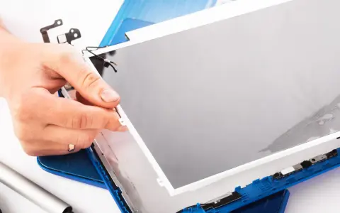 Dłoń trzyma matrycę w laptopie podczas naprawy. Widać narzędzia i niebieską obudowę.