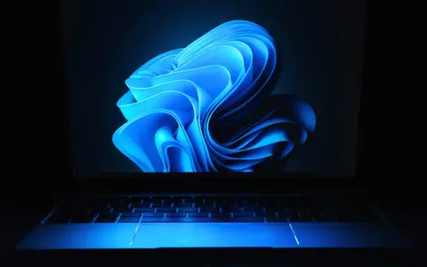 Niebieski, falujący kształt na ekranie laptopa z podświetlaną klawiaturą, przypominający logo Windows 11 24H2.