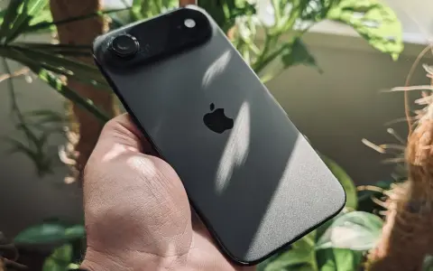 Ciemny iPhone z jednym aparatem w dłoni, otoczony zielenią roślin.