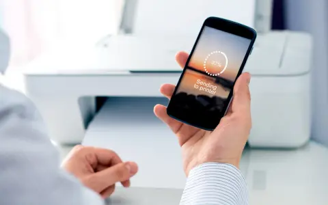 Dłoń trzyma smartfon z napisem "Sending to printer", pokazując jak drukować z telefonu. W tle biała drukarka.