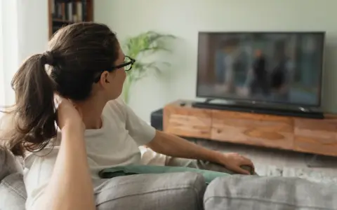 Kobieta w okularach relaksuje się na kanapie, oglądając telewizor. Idealna jaka odległość od telewizora 65 cali?