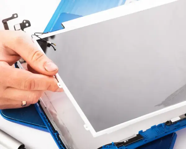 Dłoń trzyma matrycę w laptopie podczas naprawy. Widać narzędzia i niebieską obudowę.