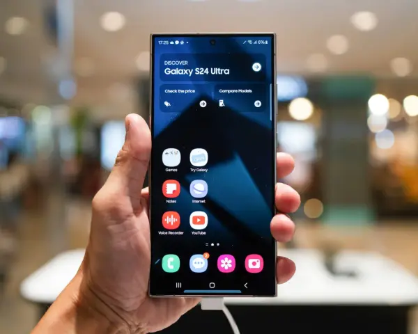 Dłoń trzyma smartfon z interfejsem One UI 6, prezentującym ekran startowy Galaxy S24 Ultra z ikonami aplikacji.
