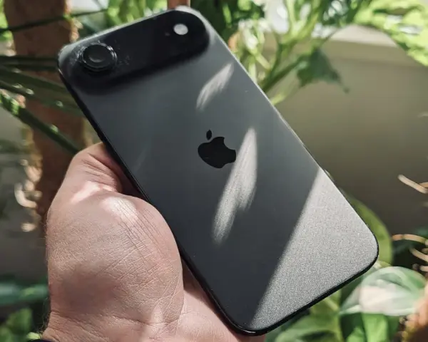 Ciemny iPhone z jednym aparatem w dłoni, otoczony zielenią roślin.