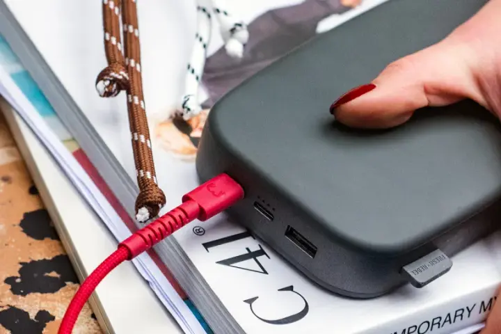 Dłoń z czerwonymi paznokciami podtrzymuje power bank 50000 mah na ile starcza, podłączony czerwonym kablem.