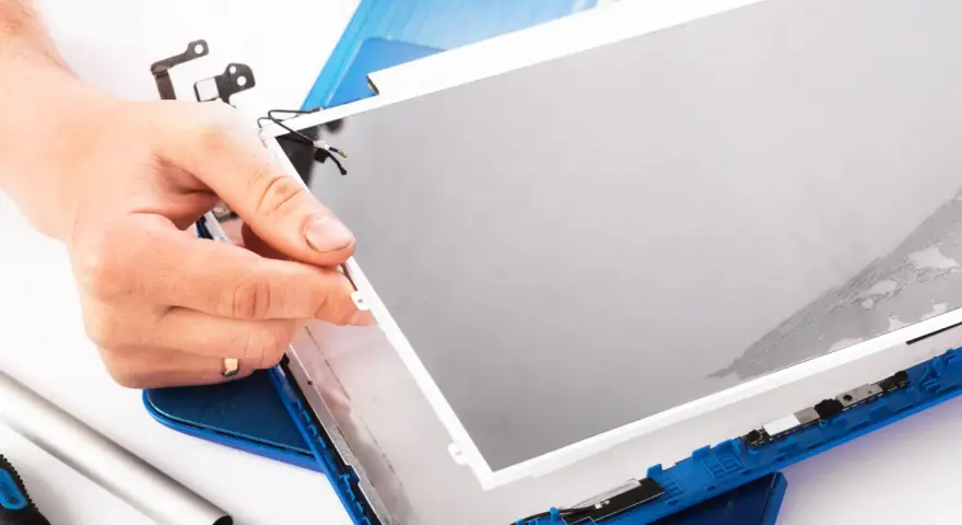 Dłoń trzyma matrycę w laptopie podczas naprawy. Widać narzędzia i niebieską obudowę.