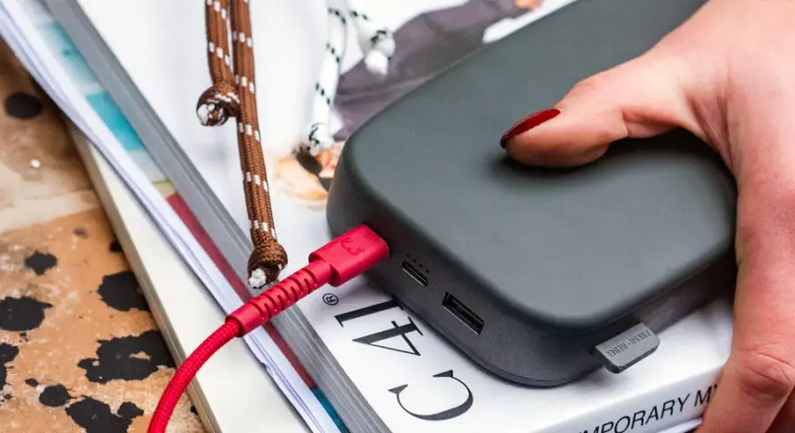 Dłoń z czerwonymi paznokciami podtrzymuje power bank 50000 mah na ile starcza, podłączony czerwonym kablem.