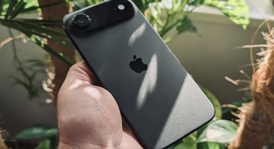Ciemny iPhone z jednym aparatem w dłoni, otoczony zielenią roślin.