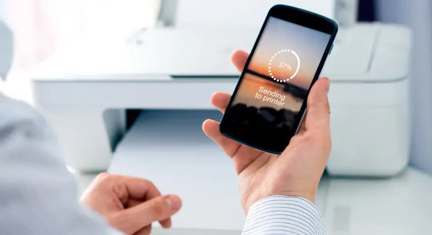 Dłoń trzyma smartfon z napisem "Sending to printer", pokazując jak drukować z telefonu. W tle biała drukarka.
