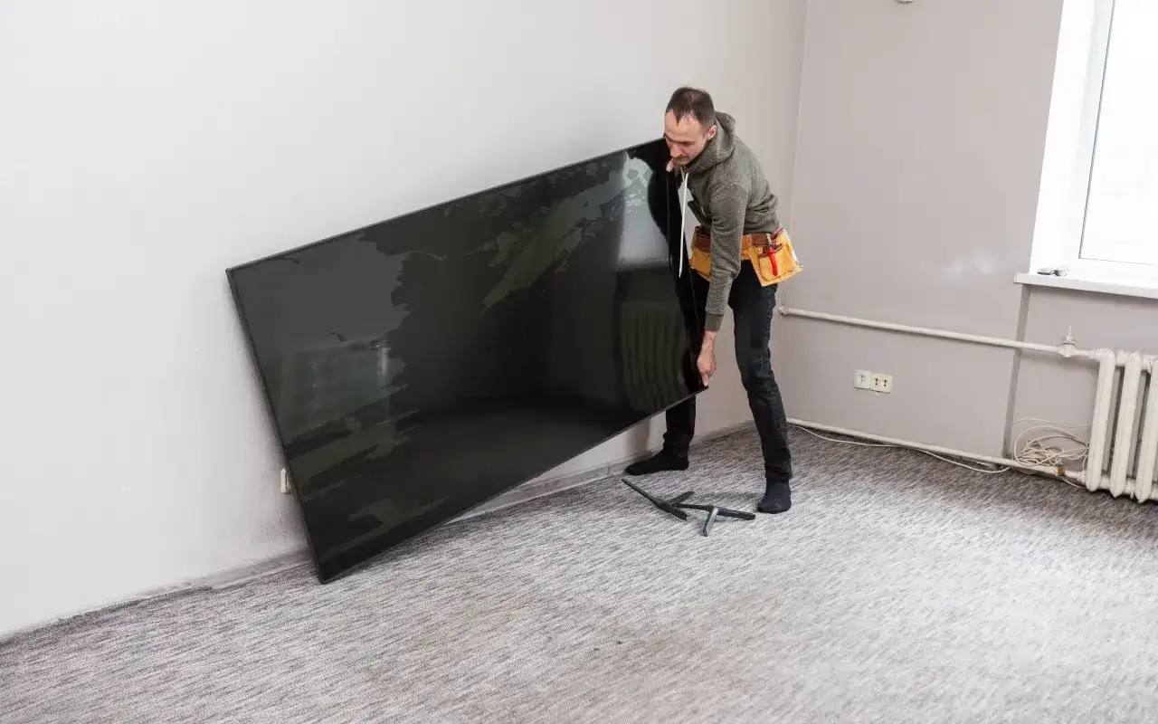 Mężczyzna z pasem narzędziowym przymierza się do powieszenia dużego telewizora. Zastanawia się, na jakiej wysokości powiesić telewizor, by był idealnie widoczny.