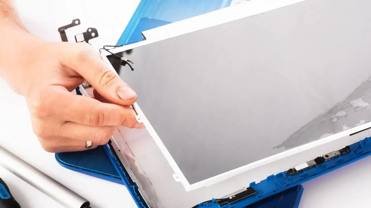 Dłoń trzyma matrycę w laptopie podczas naprawy. Widać elementy montażowe i narzędzia.