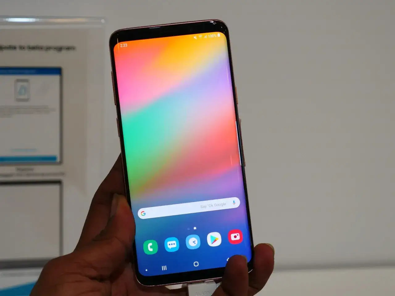 Dłoń trzyma smartfon z kolorową tapetą i ikonami aplikacji. Widoczny pasek wyszukiwania Google i przyciski nawigacyjne. To nowy interfejs One UI 8.