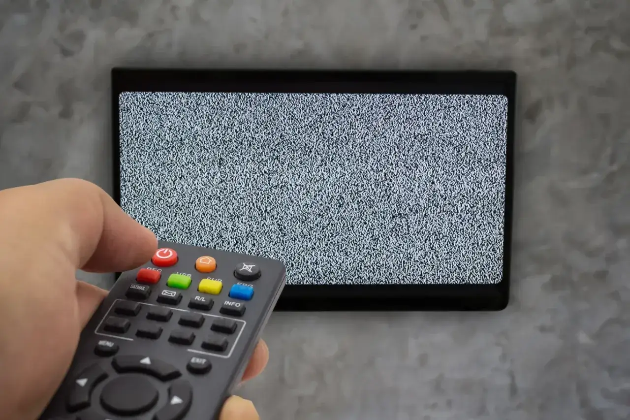 Ręka trzyma pilot dekodera, celując w ekran telewizora z zakłóceniami. Czas na sparowanie pilota z telewizorem!