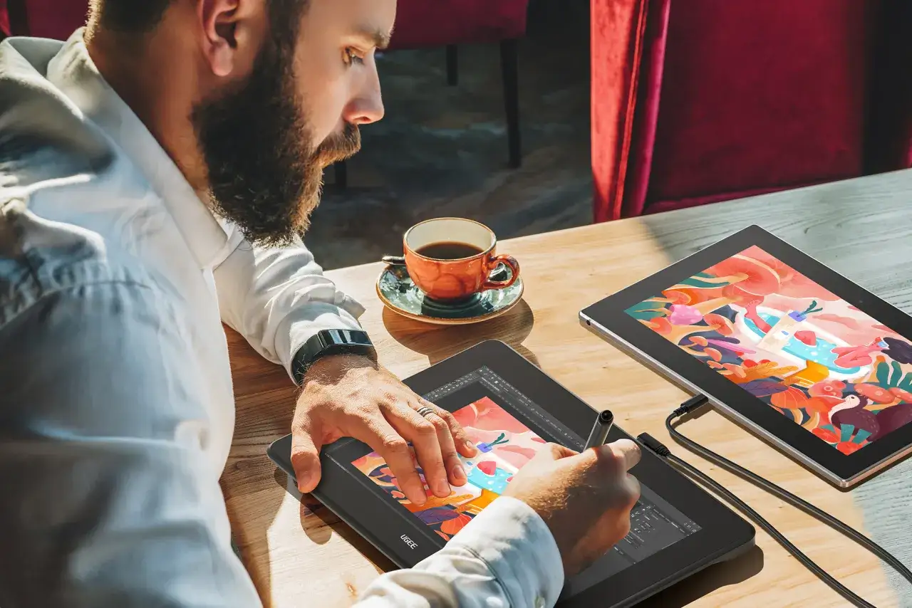 Mężczyzna z brodą tworzy cyfrowy obraz na tablecie graficznym, używając piórka. Obok stoi filiżanka kawy i drugi tablet z kolorową grafiką.